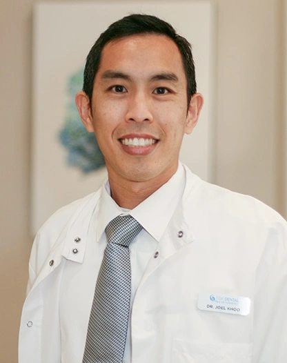 Male dental professional in white coat and tie smiling at camera in clinical setting.
