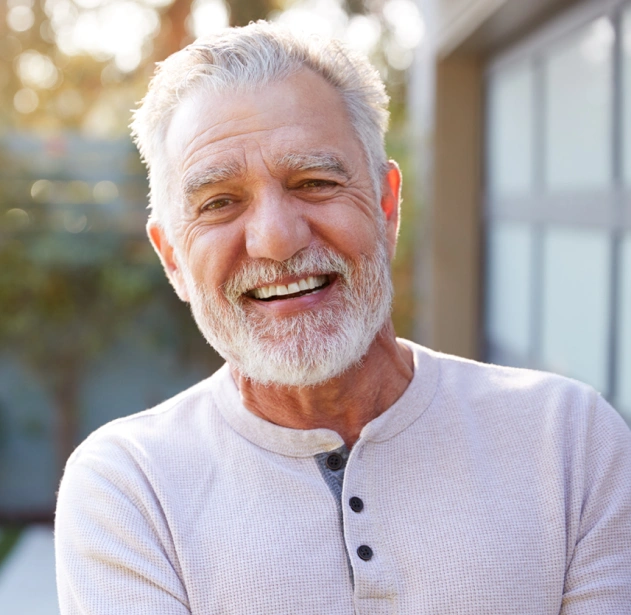 Smiling older man with white beard wearing light purple shirt outdoors near building.